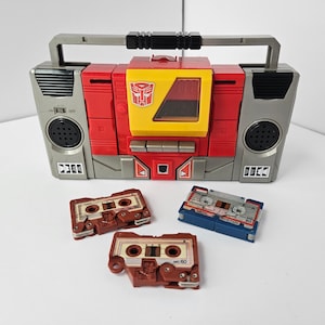 May include: A red, yellow, and gray toy boombox with a handle, speakers, and a Transformers logo. Three cassette tapes are in front of the boombox. The tapes are brown and blue.
