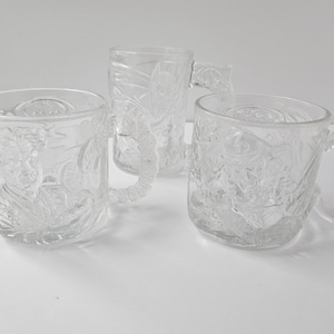 May include: Three clear glass mugs with embossed designs. Each mug has a handle and a detailed relief pattern. The mugs are arranged on a white surface, showcasing their transparent quality and intricate detailing.