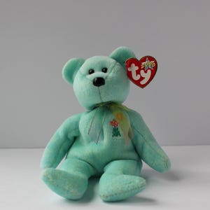 May include: A teal blue Ty Beanie Baby bear with a flower embroidered on its chest. The bear has a red heart-shaped tag with the Ty logo and the year 2000.