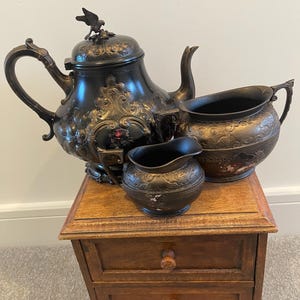 May include: A vintage tea set comprising a teapot, creamer, and sugar bowl. The dark metal set features detailed floral and scroll patterns. The teapot is topped with a bird ornament. The set is displayed on a small wooden cabinet with two drawers.
