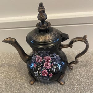 Puede incluir: Una tetera negra decorativa con detalles en bronce y un diseño floral de rosas. La tetera presenta un pico curvo, un asa y una tapa decorativa. La tetera mide aproximadamente 20 cm de altura.
