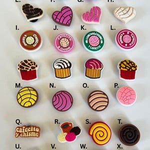 May include: A collection of colorful shoe charms featuring various designs, including pastries, hearts, and coffee-related graphics. The charms are in various shapes and colors, with text such as "Cafecito" and "Chisme".