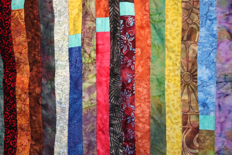 K&ouml;nnte beinhalten: Eine bunte Stoffdecke mit vertikalen Streifen in verschiedenen Rot-, Orange-, Gelb-, Gr&uuml;n-, Blau- und Lilat&ouml;nen. Der Stoff ist mit Spiralen, Blumen und geometrischen Mustern gemustert.