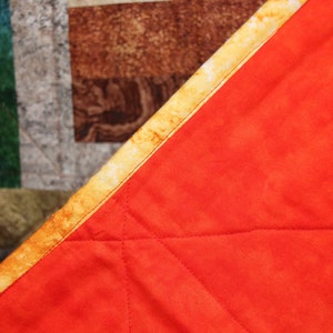 Puede incluir: Primer plano de una colcha roja con un borde estampado en amarillo y marr&oacute;n. La colcha est&aacute; doblada por la mitad y se ve la costura.