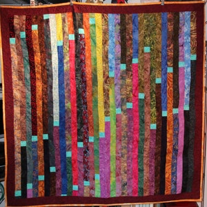 Könnte beinhalten: Ein farbenfroher Quilt aus Stoffstreifen in verschiedenen Rot-, Orange-, Gelb-, Grün-, Blau-, Lila- und Brauntönen. Die Streifen sind vertikal angeordnet und zu einem Patchwork-Muster zusammengenäht.