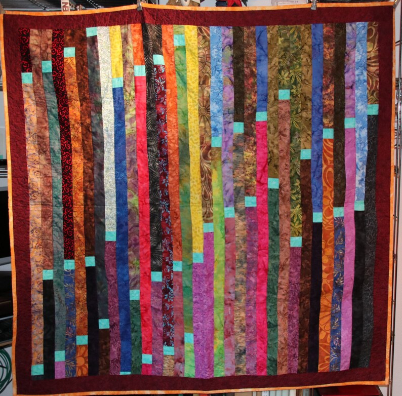 K&ouml;nnte beinhalten: Ein farbenfroher Quilt aus Stoffstreifen in verschiedenen Rot-, Orange-, Gelb-, Gr&uuml;n-, Blau-, Lila- und Braunt&ouml;nen. Die Streifen sind vertikal angeordnet und zu einem Patchwork-Muster zusammengen&auml;ht.