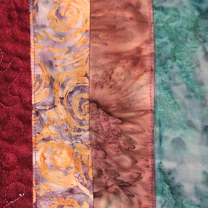 K&ouml;nnte beinhalten: Eine Nahaufnahme einer bunten Stoffdecke mit verschiedenen Mustern und Farben. Die Decke besteht aus f&uuml;nf verschiedenen Stoffstreifen, jeder mit einem einzigartigen Design. Der Stoff links ist tief burgunderfarben mit einem gesteppten Muster. Der n&auml;chste Streifen ist ein hellvioletter und gelber Stoff mit einem wirbelnden Muster. Der dritte Streifen ist ein hellbrauner und rosa Stoff mit einem Batik-Muster. Der vierte Streifen ist ein hellblauer und gr&uuml;ner Stoff mit einem Batik-Muster. Der letzte Streifen ist ein schwarzer Stoff mit einem roten Wirbelmuster.