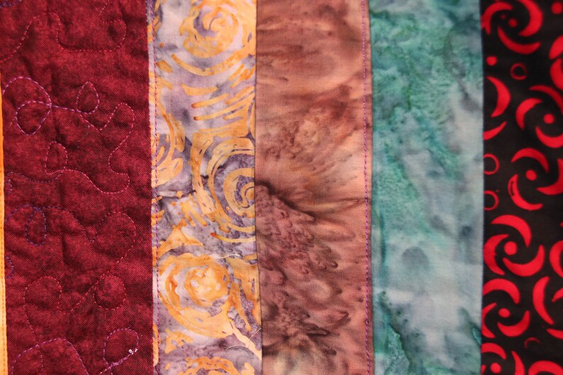 K&ouml;nnte beinhalten: Eine Nahaufnahme einer bunten Stoffdecke mit verschiedenen Mustern und Farben. Die Decke besteht aus f&uuml;nf verschiedenen Stoffstreifen, jeder mit einem einzigartigen Design. Der Stoff links ist tief burgunderfarben mit einem gesteppten Muster. Der n&auml;chste Streifen ist ein hellvioletter und gelber Stoff mit einem wirbelnden Muster. Der dritte Streifen ist ein hellbrauner und rosa Stoff mit einem Batik-Muster. Der vierte Streifen ist ein hellblauer und gr&uuml;ner Stoff mit einem Batik-Muster. Der letzte Streifen ist ein schwarzer Stoff mit einem roten Wirbelmuster.