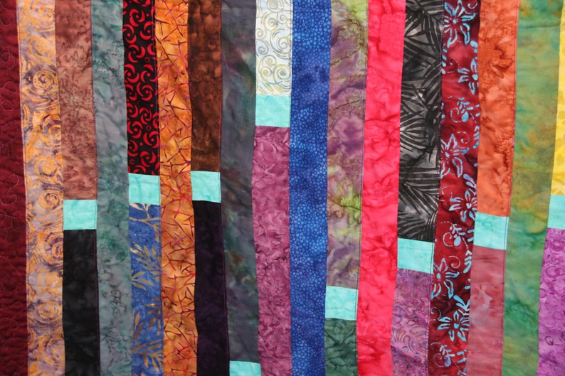 K&ouml;nnte beinhalten: Ein farbenfroher Quilt aus Stoffstreifen in verschiedenen Mustern und Farben, darunter Rot, Orange, Gelb, Gr&uuml;n, Blau, Lila und Braun. Der Quilt besteht aus vertikalen Stoffstreifen, wobei einige horizontale Stoffstreifen dazwischen eingen&auml;ht sind.