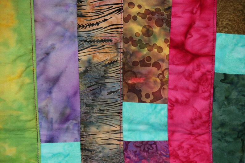 K&ouml;nnte beinhalten: Ein Patchwork-Deckenst&uuml;ck mit verschiedenen Farben und Mustern, darunter Gr&uuml;n, Lila, Braun, Rosa und T&uuml;rkis. Die Decke besteht aus verschiedenen Stoffen, die zusammengen&auml;ht wurden, um ein einzigartiges Design zu schaffen.