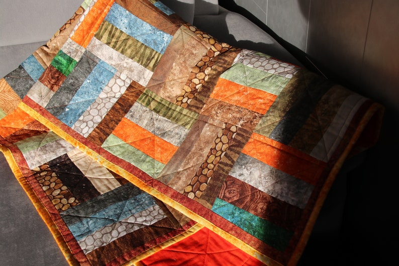 Puede incluir: Una colcha de retazos con un patr&oacute;n geom&eacute;trico en tonos marrones, naranjas, verdes y azules. La colcha est&aacute; hecha de diferentes telas con diversas texturas, incluyendo una tela con un patr&oacute;n de guijarros.