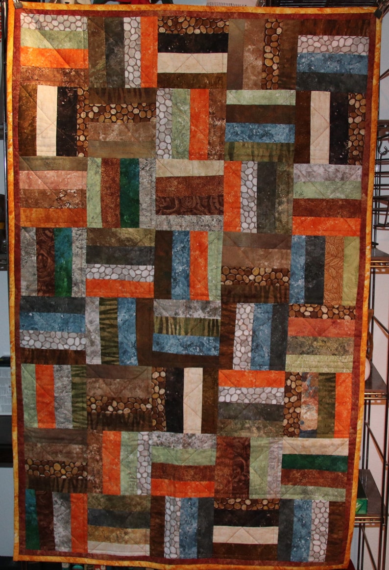 Puede incluir: Una colcha hecha de varios retazos de tela en tonos marrones, naranjas, verdes y azules. La colcha est&aacute; hecha de tiras horizontales y verticales de tela, creando un dise&ntilde;o de patchwork.