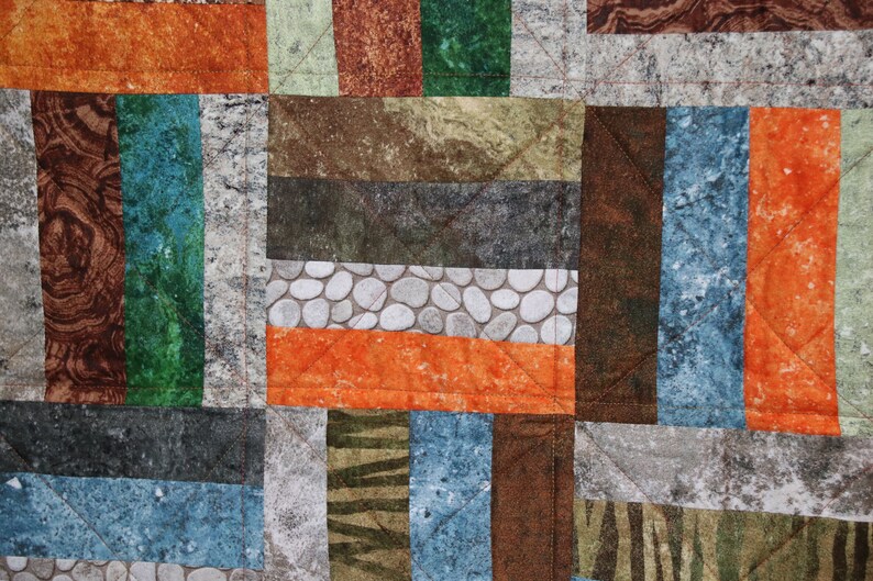 Puede incluir: Una colcha de retazos con una variedad de telas marrones, naranjas, verdes y azules. Las telas tienen diferentes patrones, incluyendo piedras, rayas y veta de madera.