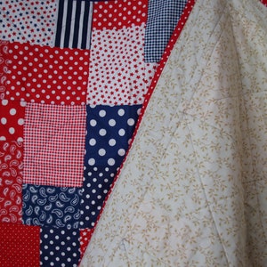 Puede incluir: Una colcha de retazos rojo, blanco y azul con un reverso blanco con un peque&ntilde;o estampado floral. La colcha est&aacute; hecha de varias telas con diferentes patrones, incluyendo lunares, estrellas y paisley.