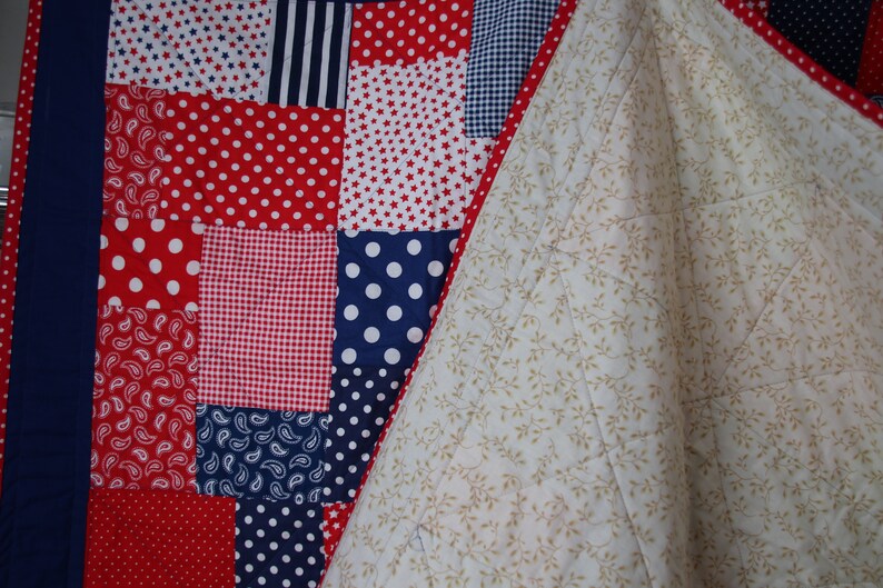 Puede incluir: Una colcha de retazos rojo, blanco y azul con un reverso blanco con un peque&ntilde;o estampado floral. La colcha est&aacute; hecha de varias telas con diferentes patrones, incluyendo lunares, estrellas y paisley.