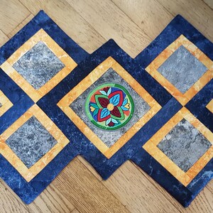 Könnte beinhalten: Ein blauer und gelber gesteppter Tischläufer mit fünf gestickten Blumendesigns. Der Läufer ist aus Stoff mit einem marmorartigen Muster.