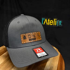 Peut inclure: Casquette de camionneur grise avec un dos en filet noir et un patch en cuir marron. Le patch présente des icônes sur le thème du camping et le mot "REPEAT". La casquette a une visière incurvée et une étiquette "Richardson".