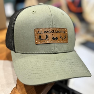 Peut inclure: Casquette de camionneur vert olive et noir avec une visière incurvée. Le chapeau comporte un écusson en cuir marron avec le texte "ALL RACKS MATTER" et des silhouettes de bois de cerf, d'un canard et d'autres bois de cerf. Le chapeau est en tissu durable.
