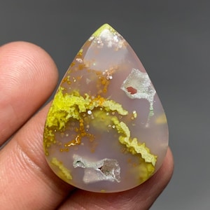 May include: A teardrop-shaped polished gemstone featuring a translucent pale lavender base with vibrant yellow moss-like inclusions, scattered orange spots, and small white crystalline vugs embedded within the smooth, rounded surface.