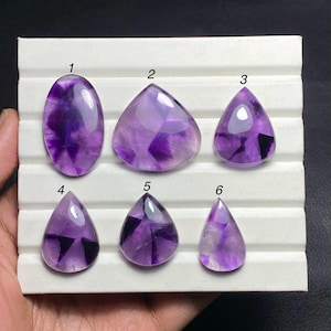 Pode incluir: Seis pedras preciosas de ametista polidas em várias formas, incluindo oval, triangular e em forma de gota. As pedras exibem uma gama de tons roxos com inclusões mais escuras, sobre um fundo branco.