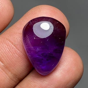 May include: A polished, teardrop-shaped amethyst gemstone. The stone transitions from a deep purple at the base to a lighter, translucent purple at the top, with a white, oval-shaped inclusion near the top. The gemstone is held in a person's fingers.