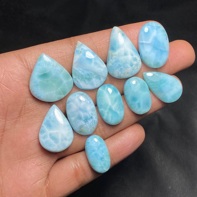 May include: A collection of polished larimar gemstones in various shapes, including teardrop and oval. The stones display a range of light blue hues with white marbling, resembling ocean waves. The gemstones are held in a hand against a dark background.