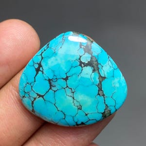 Puede incluir: Una piedra preciosa turquesa pulida, de forma triangular, de color azul vibrante. La piedra presenta una compleja matriz de vetas marrón oscuro, creando un patrón único. La piedra preciosa se sostiene en una mano, mostrando su tamaño y forma.