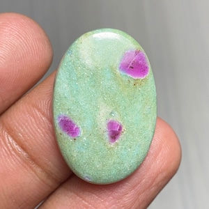 Puede incluir: Gema ovalada con una base de color verde claro y varias inclusiones rosas vibrantes. La piedra pulida tiene una superficie lisa y una forma ligeramente irregular, mostrando variaciones naturales de color y textura. La gema se sostiene en una mano.