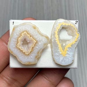 May include: Two irregular agate slices labeled 1 and 2. The left slice features concentric white and cream bands surrounding a crystalline center. The right slice has a translucent white outer edge with a bright yellow inner ring and a hollow center.