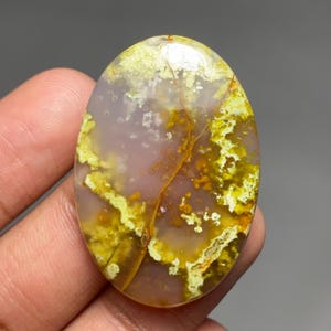 May include: An oval-shaped cabochon gemstone featuring a translucent, pale lavender base colour with intricate, mossy inclusions in shades of chartreuse, mustard yellow, and deep amber, creating a natural, organic pattern across the smooth surface.