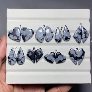 May include: A collection of eight pairs of snowflake obsidian cabochons in various shapes, including hearts, teardrops, and geometric forms. The stones feature a black base with white snowflake-like patterns. The cabochons are displayed on a white tray.