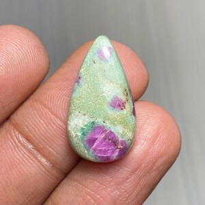 Puede incluir: Una gema en forma de lágrima con una matriz verde claro, salpicada de inclusiones de rubí rosa y rojo vibrantes. La piedra pulida se sostiene en una mano, mostrando su belleza natural y variaciones de color únicas. Ideal para la fabricación de joyas.