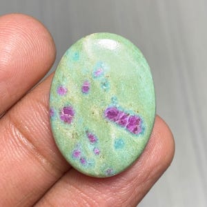 Puede incluir: Gema ovalada con un color base verde claro, salpicada de inclusiones rosas y turquesas vibrantes. La superficie pulida de la piedra refleja la luz, resaltando los patrones y texturas naturales. Ideal para joyería o coleccionismo.