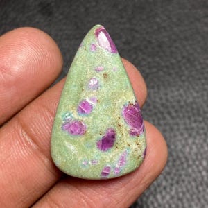 Op de afbeelding: Een peervormige cabochon edelsteen met een lichtgroene zoisiet matrix met ingebedde levendige magenta-roze robijninsluitingen. De steen heeft een glad, gepolijst oppervlak met een druppelvormig silhouet en zichtbare natuurlijke mineraalvariaties.