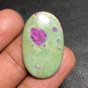 Op de afbeelding: Een ovaalvormige cabochon edelsteen met een gladde, lichtgroene basiskleur, een opvallende, onregelmatige magenta-roze insluiting nabij het midden en verschillende kleinere, verspreide magenta-roze vlekken over het oppervlak.