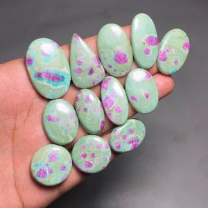 May include: A collection of eleven polished cabochon gemstones featuring a light mint green base colour with vibrant magenta and pink inclusions. These smooth, rounded stones exhibit a glossy finish and varied oval and teardrop shapes.