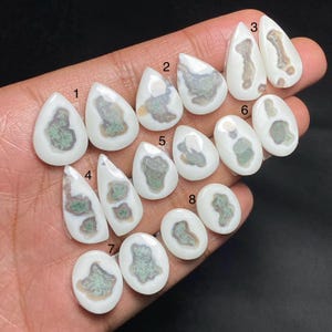 May include: A collection of polished gemstones in various shapes, including teardrop and oval. The stones feature a creamy white base with intricate patterns of green and brown, creating a unique and natural aesthetic. The gemstones are held in a hand.