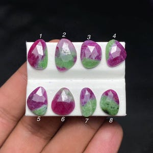 Può includere: Otto pietre preziose sfaccettate di rubino e zoisite disposte in due file su un cartoncino bianco. Ogni pietra presenta una combinazione unica di colori rosa magenta vibrante e verde muschio. Le pietre sono numerate da uno a otto con inchiostro nero.