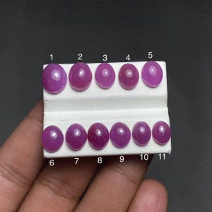 May include: Eleven oval-shaped, smooth, magenta-colored cabochon gemstones arranged in two rows on a white display stand. Each stone is numbered from 1 to 11 in black text above or below the corresponding gem, showcasing a polished, reflective surface.