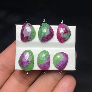 May include: Six faceted ruby zoisite gemstones arranged in two rows on a white display card. Each stone features a unique blend of vibrant magenta and deep green colours with numbered labels from one to six positioned above or below each gem.