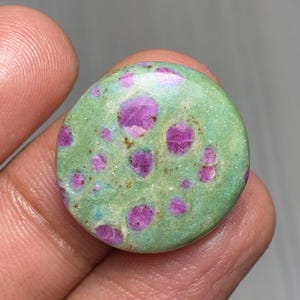 Puede incluir: Una gema redonda y pulida con una base de color verde claro. La piedra presenta numerosas manchas rosas vibrantes dispersas por su superficie. La gema se sostiene en una mano, mostrando su tamaño y textura. La apariencia general sugiere un origen natural y orgánico.