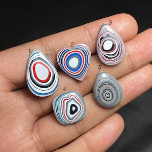 May include: Five polished stones with a distinctive striped pattern in red, white, black, and grey. The stones are in various shapes, including teardrop, heart, and oval. The stones are held in a hand.