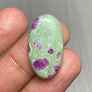 Puede incluir: Una gema ovalada con una base verde claro e inclusiones magenta. La piedra pulida presenta un patrón moteado de tonos magenta, creando una apariencia única. La gema se sostiene en una mano. La piedra mide aproximadamente 3 cm de largo.