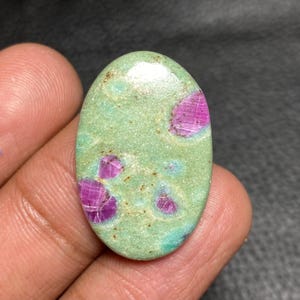 Op de afbeelding: Een ovaalvormige cabochon edelsteen met een gladde, lichtgroene basis en opvallende, ingebedde magenta-kleurige insluitingen. Het oppervlak heeft een gepolijste, reflecterende afwerking met subtiele natuurlijke spikkels in de textuur.
