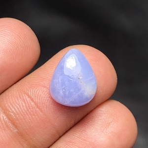 May include: A teardrop-shaped cabochon gemstone with a smooth, polished surface and a light periwinkle blue color. The stone features subtle internal white veining and a reflective luster, showcasing a translucent quality throughout the piece.