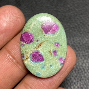 Op de afbeelding: Een ovaalvormige cabochon edelsteen met een gladde, lichtgroene basis en verschillende ingebedde, diep magenta gekleurde kristalinsluitingen. Het oppervlak heeft een gepolijste afwerking met zichtbare natuurlijke textuur en kleine bruine mineraalvlekjes.