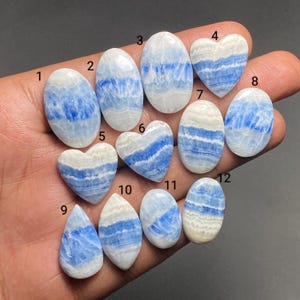 May include: Twelve polished gemstones featuring horizontal bands of white and blue, labeled with numbers one through twelve. The collection includes various shapes such as ovals, hearts, and a teardrop, all displaying a smooth, glossy surface texture.