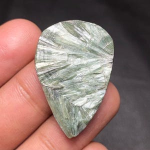 Puede incluir: Una gema pulida en forma de lágrima con una textura fibrosa única. La piedra presenta una mezcla de tonos verde pálido y blanco, con un patrón radiante que emana del centro. La gema se sostiene en una mano, mostrando su belleza natural.