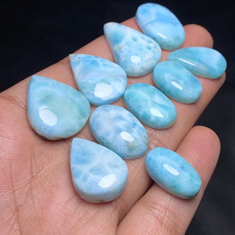 May include: A collection of polished larimar gemstones in various shapes, including teardrop and oval. The stones display a range of light blue hues with white marbling, creating a sea-like appearance. The gemstones are smooth and reflective, with a glossy finish.