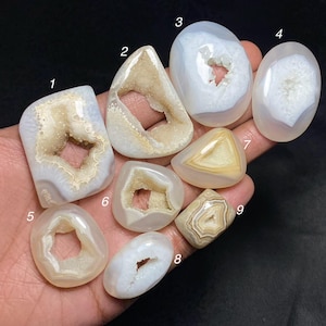 May include: A selection of polished agate geodes in diverse shapes and sizes. The geodes showcase a spectrum of colours, including white, beige, and brown, with crystalline interiors. The collection is held in a hand against a dark backdrop.